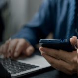 A picture of a man holding a mobile phone and on a laptop, symbolising the modernisation of trust compliance.